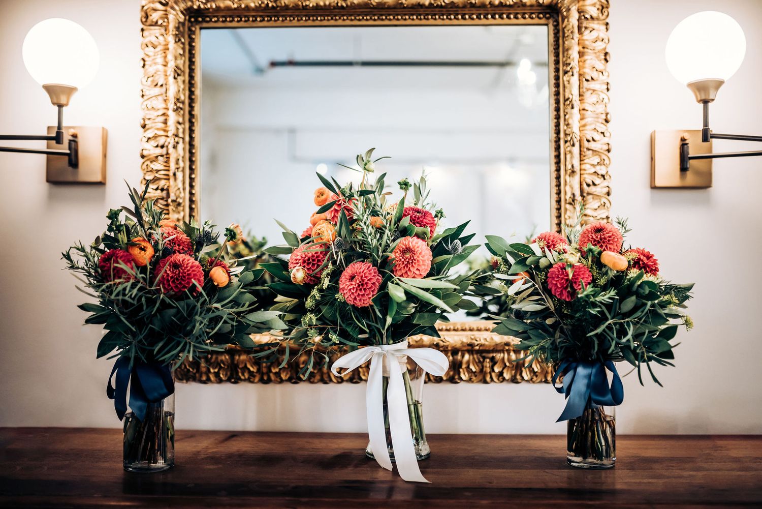 Three elegant floral arrangements with coral roses displayed on ornate gold mirror.