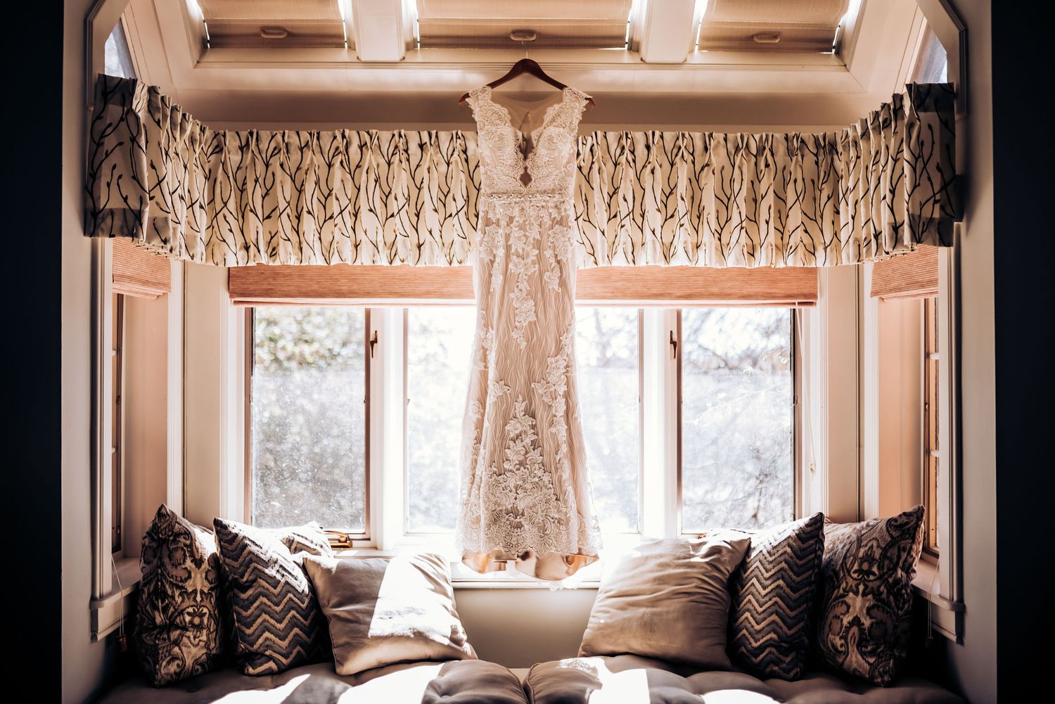 Elegant white wedding dress hangs in window nook with decorative pillows.
