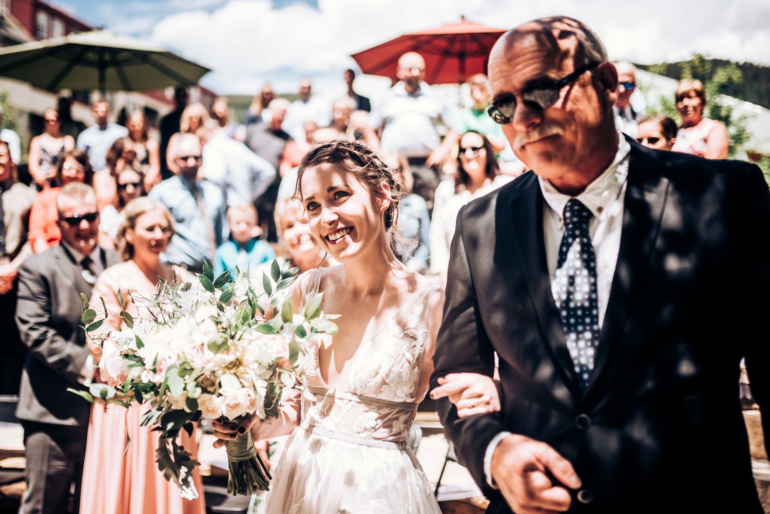 Sun-drenched outdoor wedding ceremony with crowd in background.