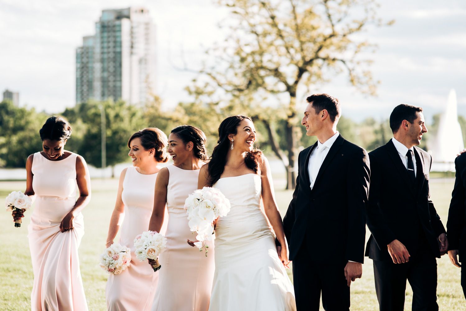 Wedding party in formal attire walking together on grassy lawn with cityscape in background.