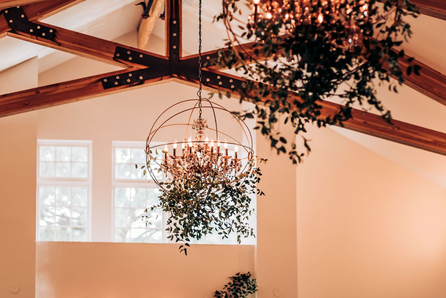 Ornate crystal chandelier hangs from wooden beams with cascading greenery in an elegant wedding venue.