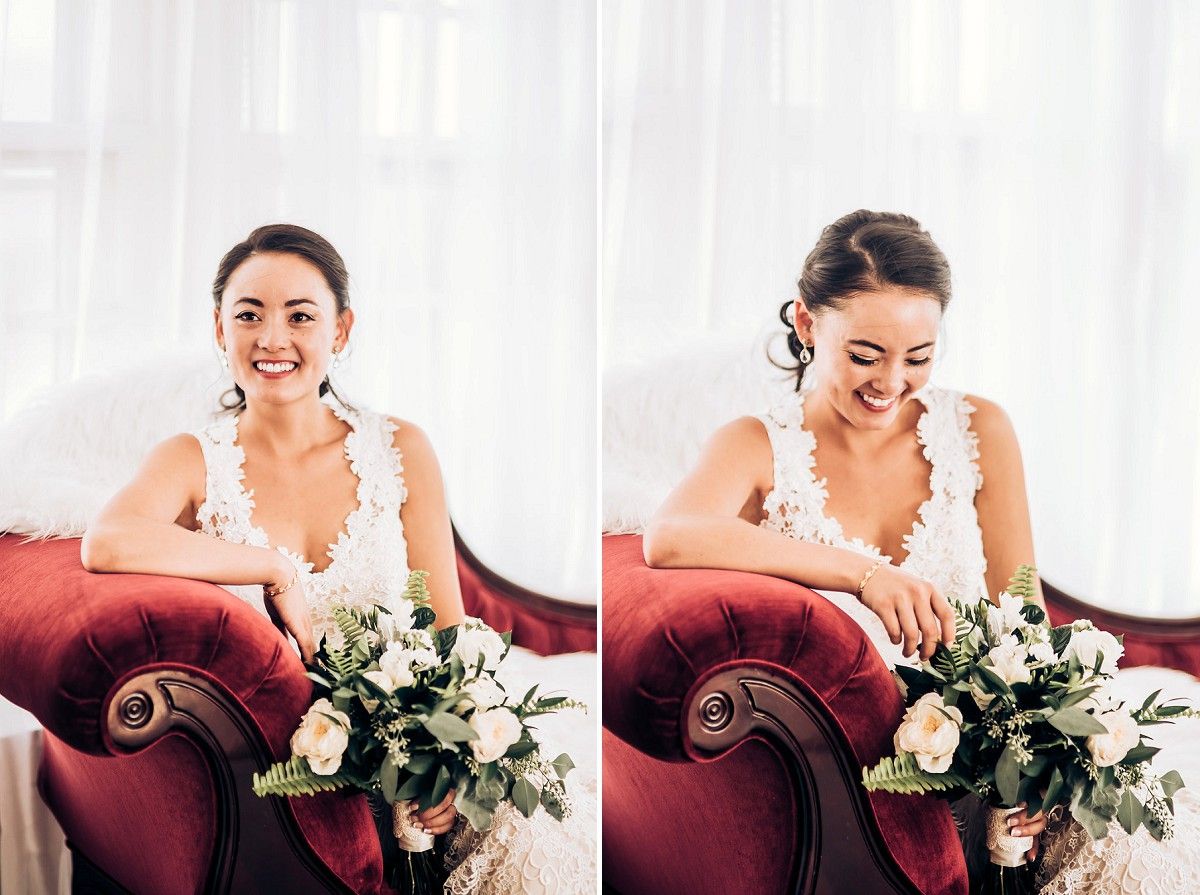 Bride with bouquet laughing while seated on red velvet couch in bright sunlit room.