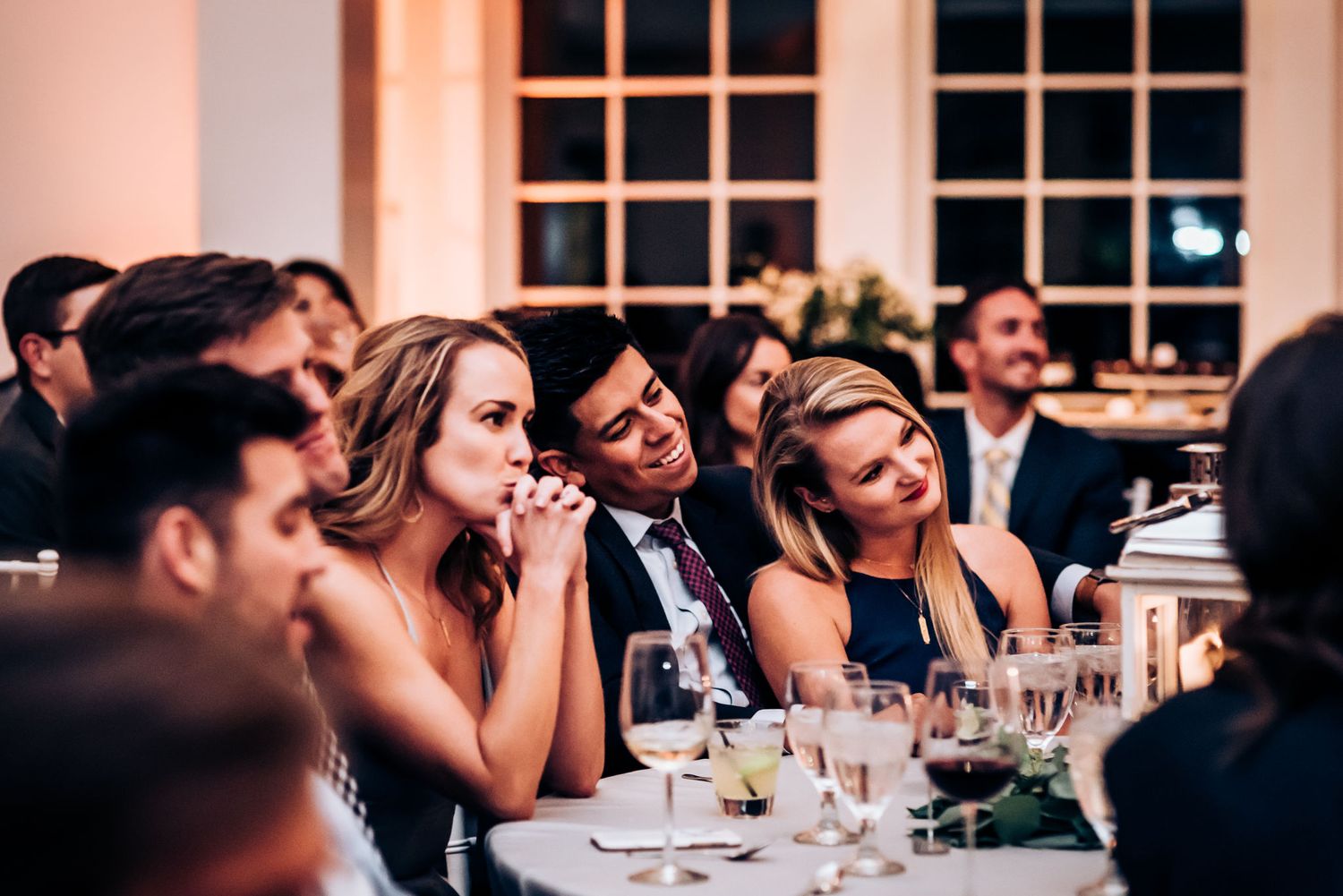 Wedding guests enjoy dinner and speeches at elegant reception with floral decor.