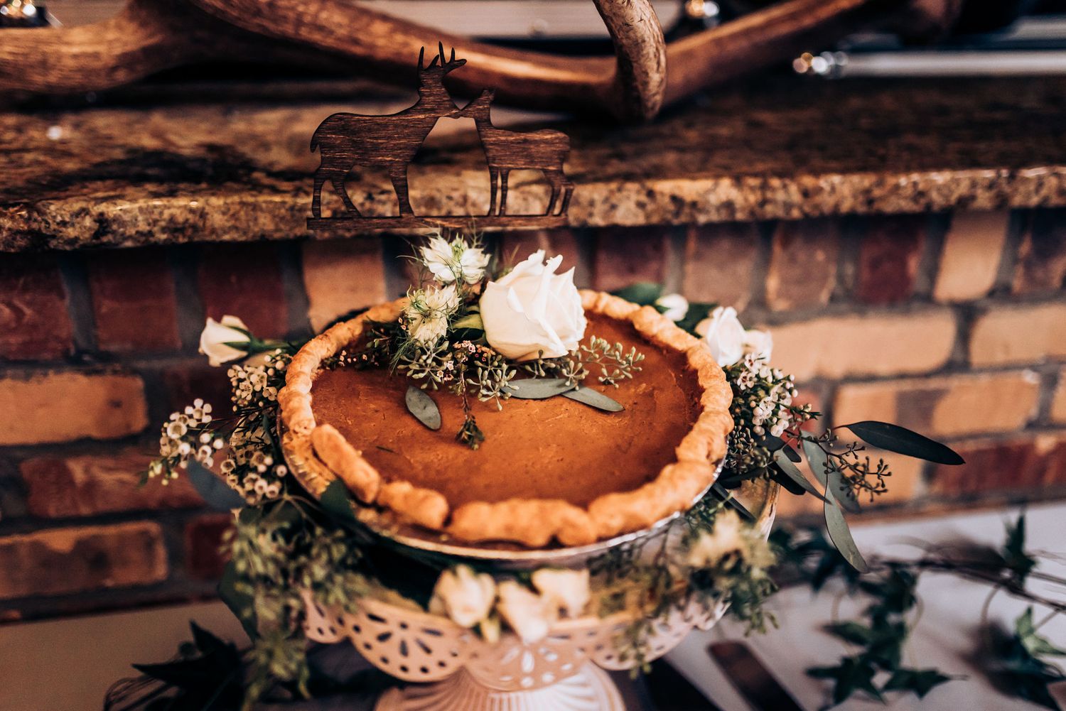 Rustic wedding dessert table featuring a pie decorated with flowers against brick wall.