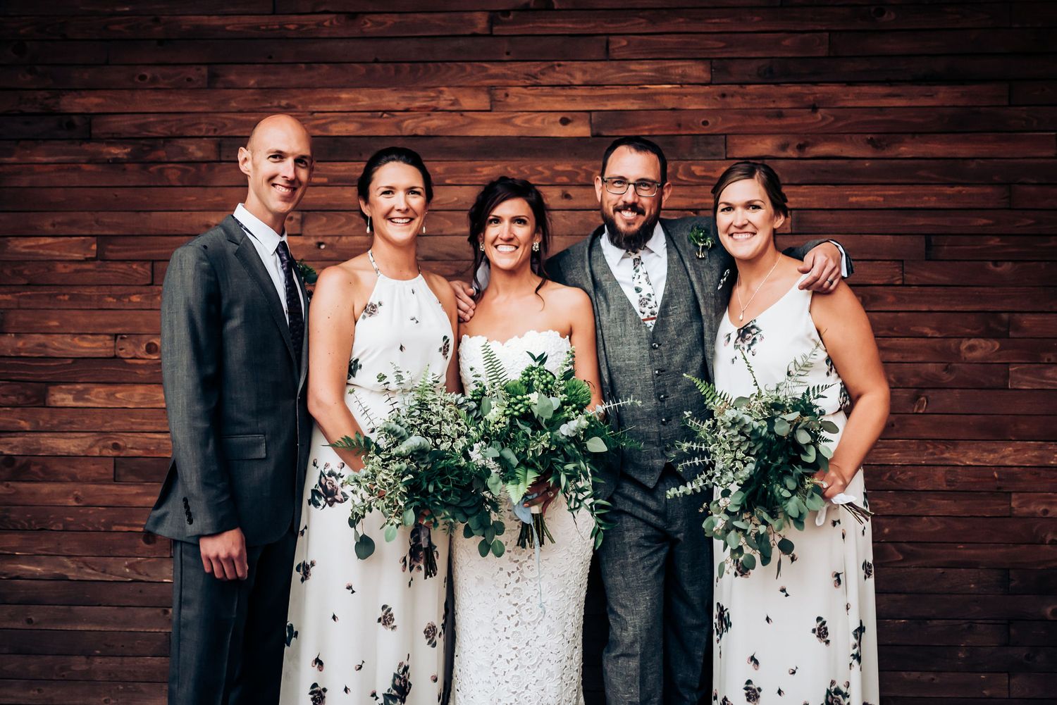 Wedding party in floral bridesmaid dresses poses against rustic wooden wall with lush greenery bouquets.