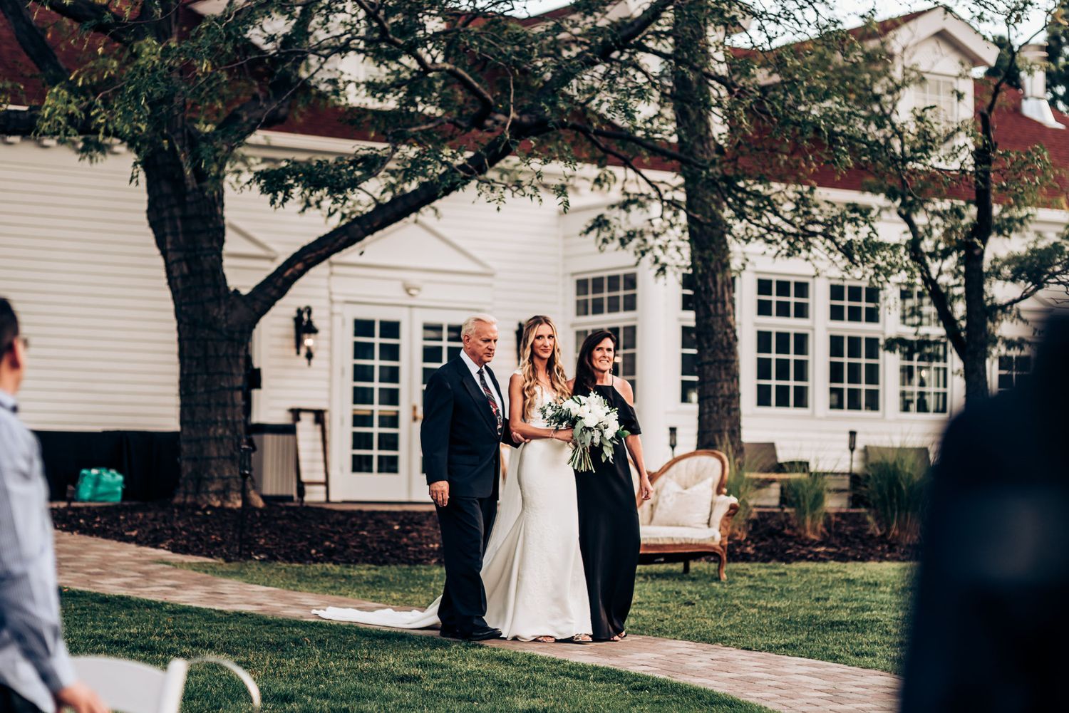 Outdoor wedding ceremony in front of white building with vintage furniture and trees.