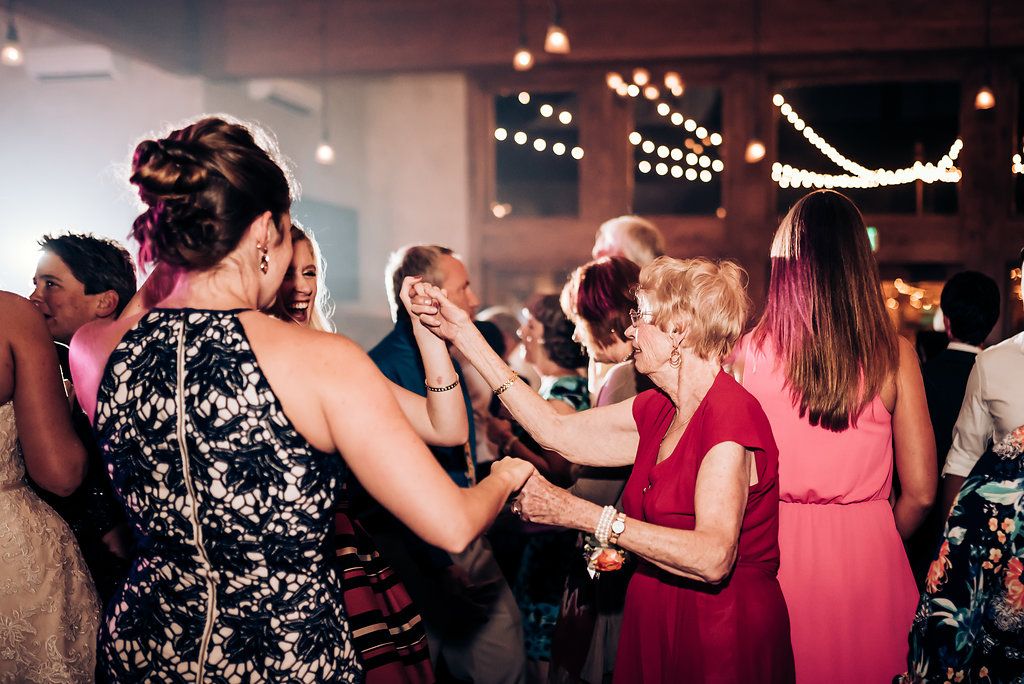 Wedding guests dancing and celebrating in formal attire under decorative string lights at an indoor reception.