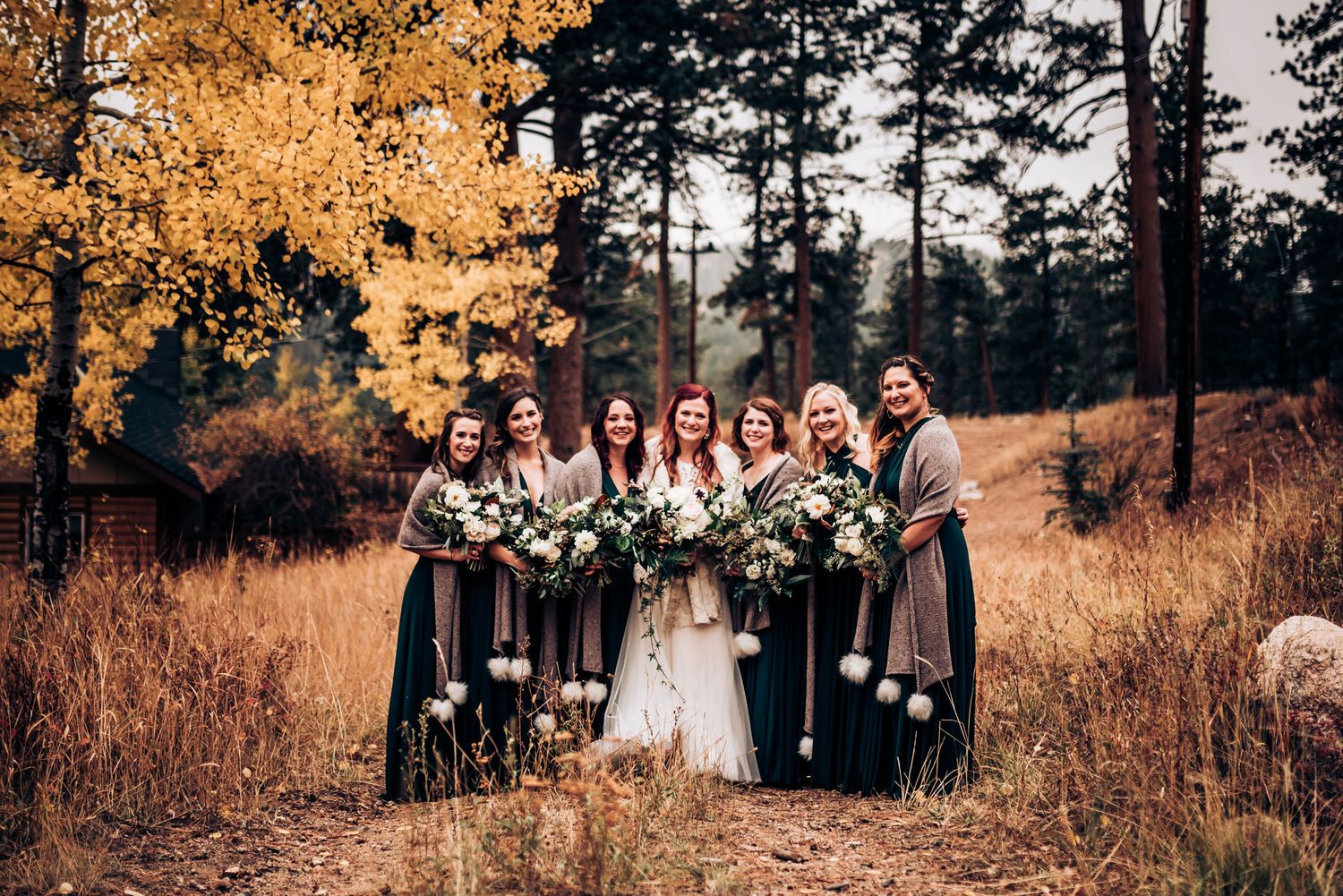 Bridal party in dark green dresses poses against autumn trees and golden grass.