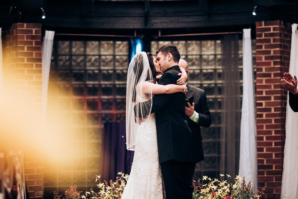 Newlyweds share their first kiss as a married couple in front of a brick wall with flowers.