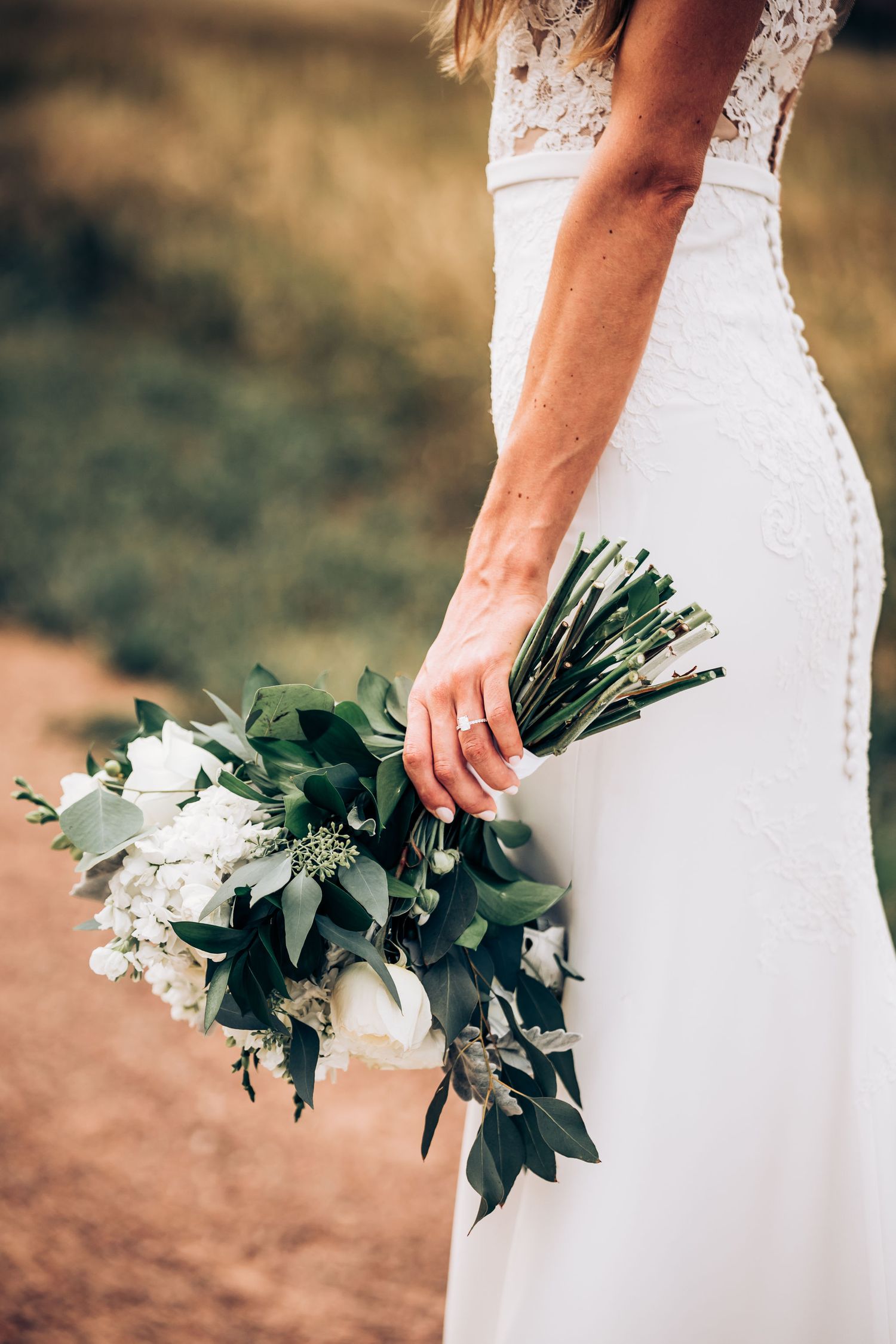 Side view of delicate white wedding bouquet with greenery held against a lace wedding gown outdoors.