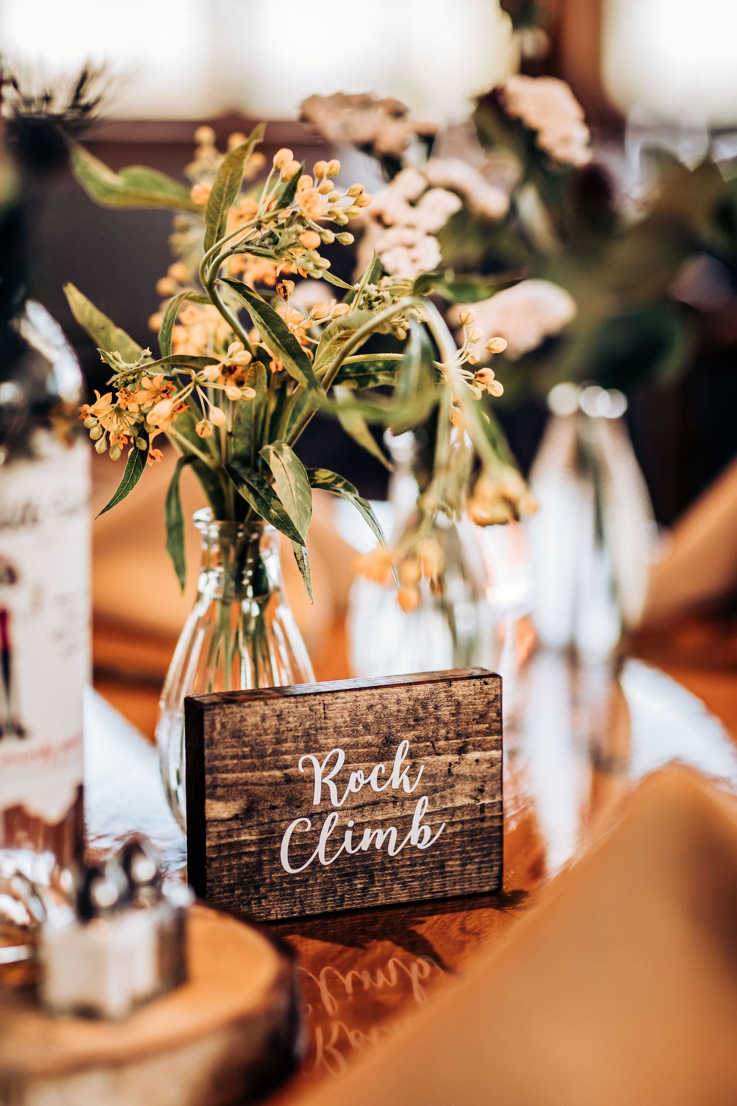 Rustic wedding table centerpiece with wildflowers in glass vase and wooden 'Rock Climb' sign.