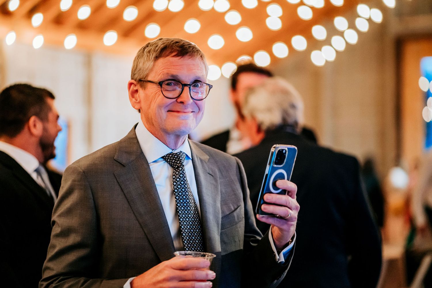 Guest in formal attire holding phone and drink under string lights at evening event.