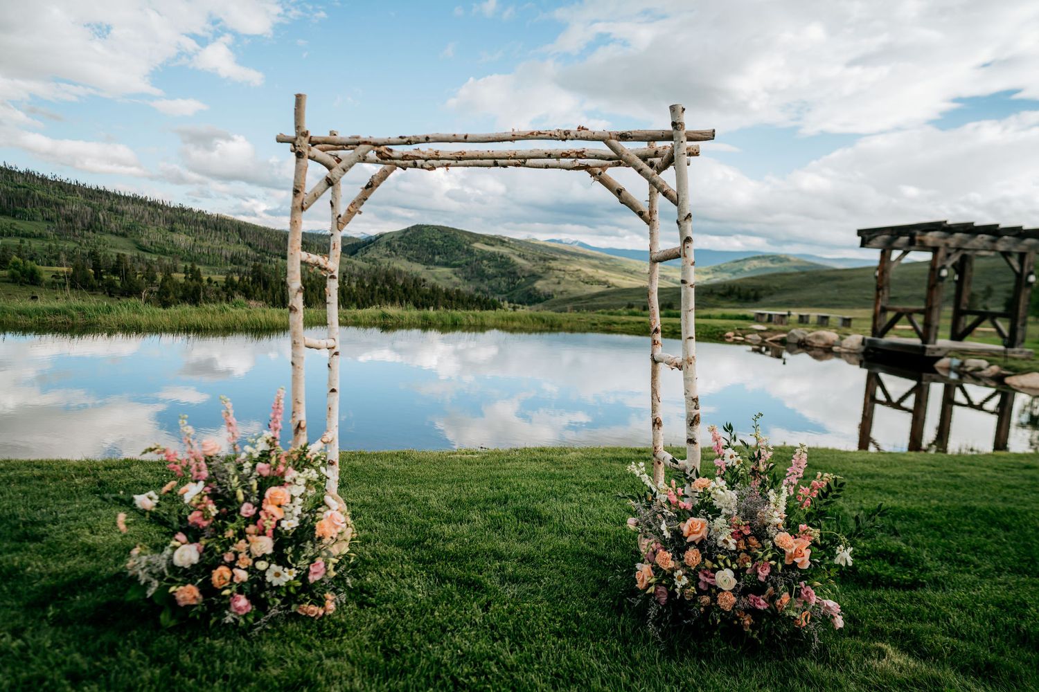 Rustic wooden wedding arch decorated with pink and peach flowers overlooking a mountain lake.