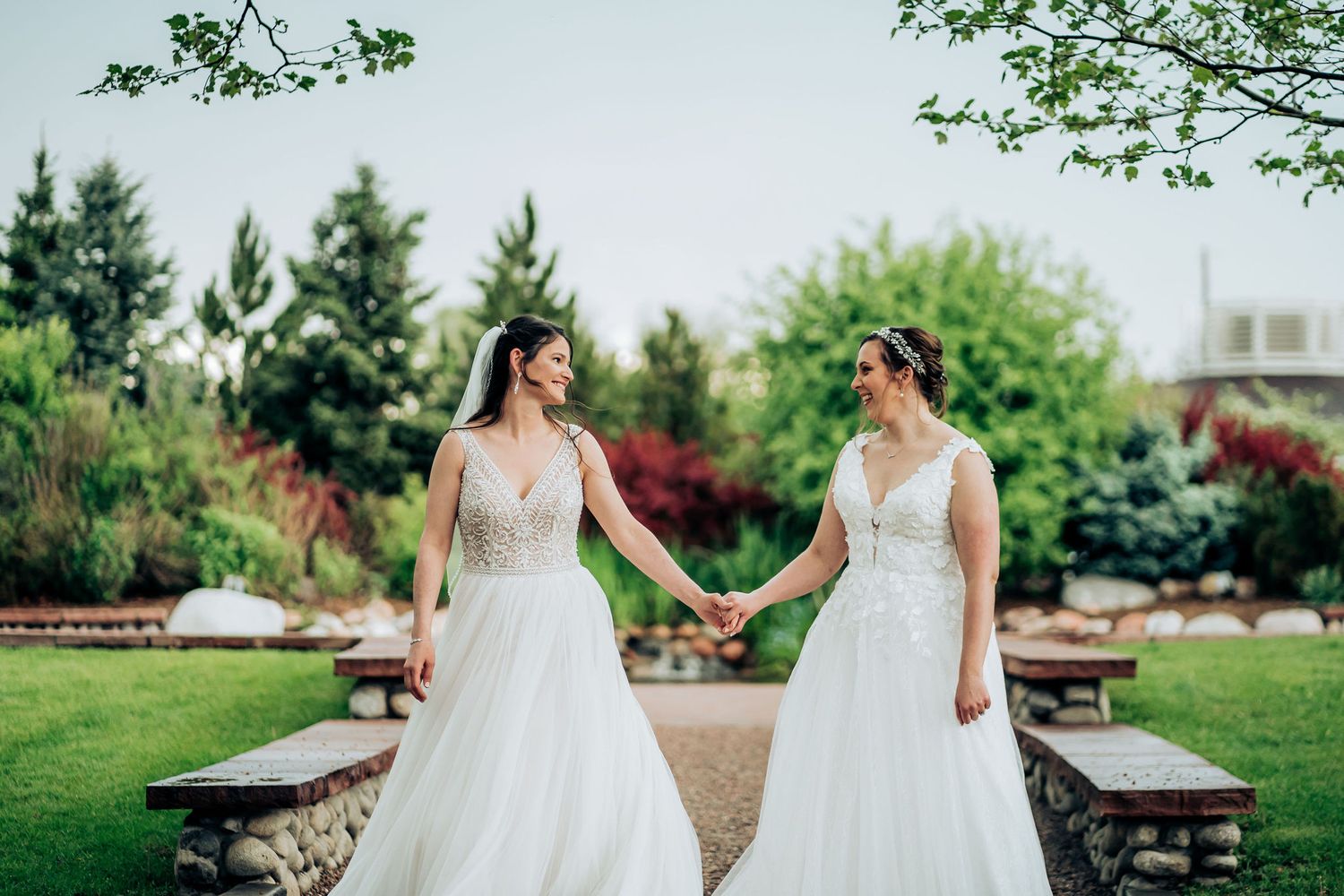 Two brides in elegant white wedding gowns hold hands in a manicured garden setting.