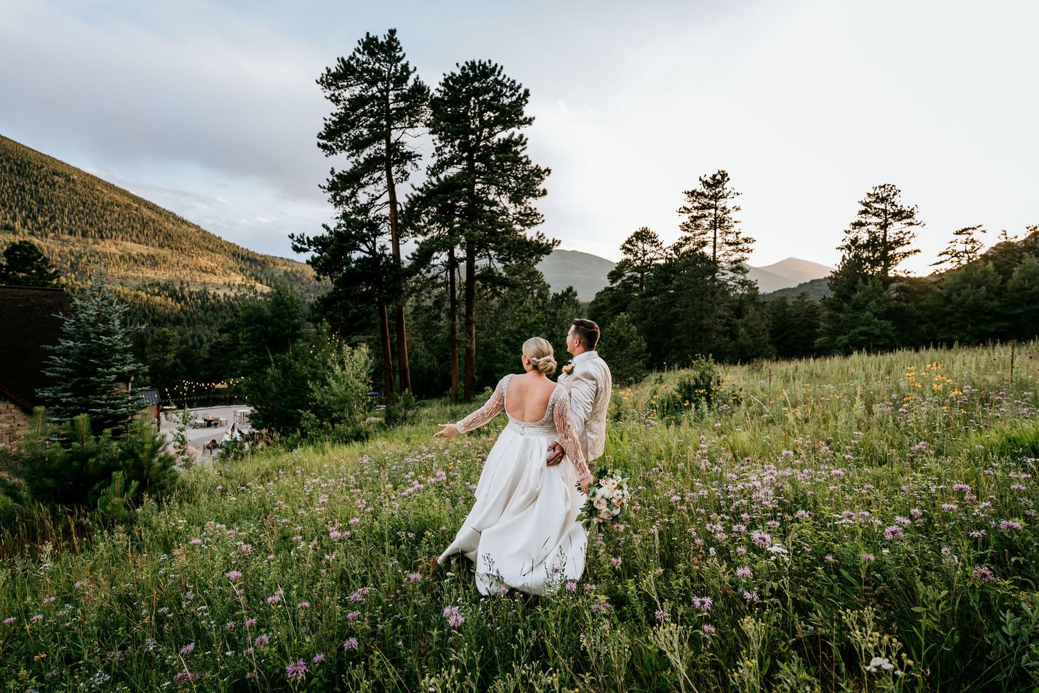Wedding couple walks through mountain wildflower meadow with pine trees in background.