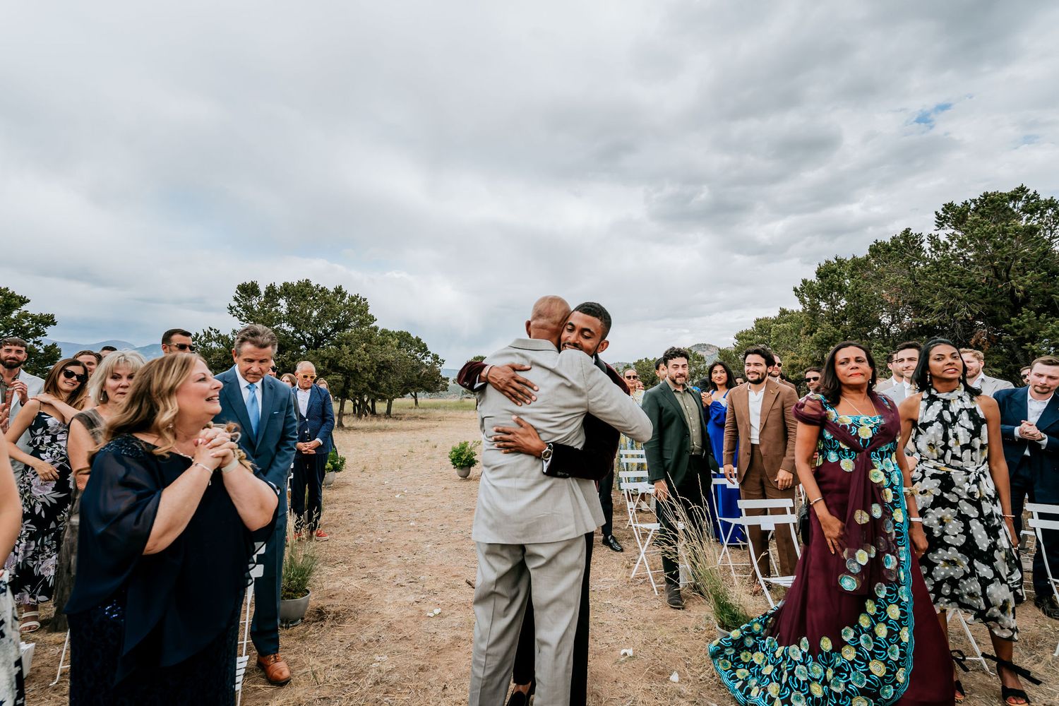 Wedding guests celebrate as a couple embraces during an outdoor ceremony.