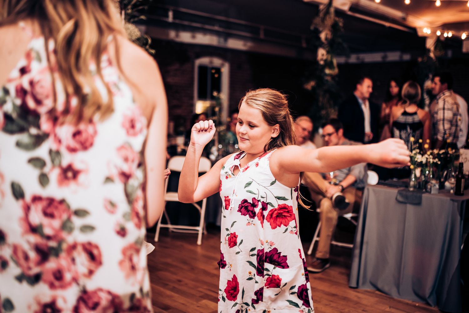 Dancers in floral dresses move energetically on a wooden dance floor at a wedding celebration.