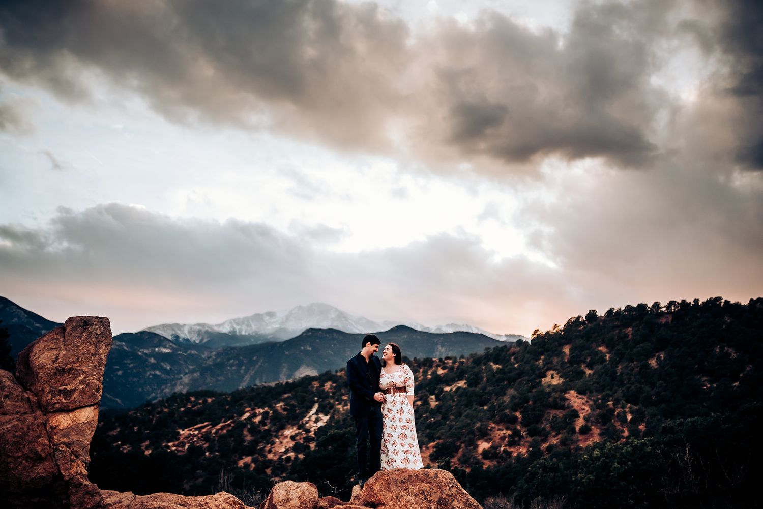 Silhouetted couple stands atop rocky mountain overlook during dramatic cloudy sunset.