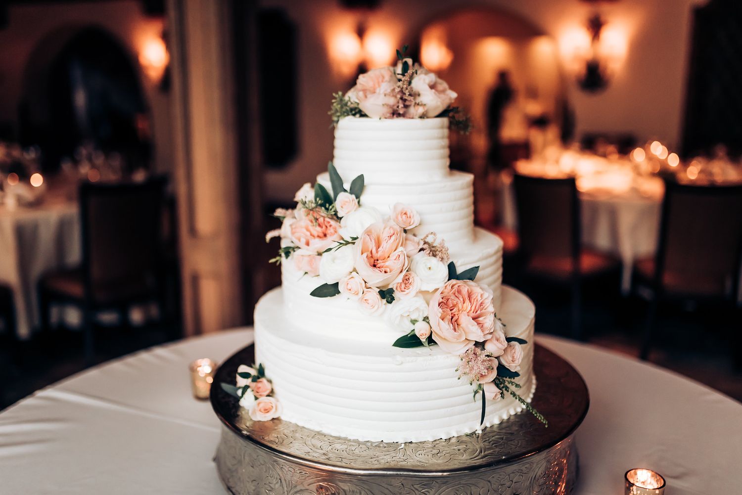 Elegant white tiered wedding cake decorated with peach-colored flowers and greenery on display at a reception venue.