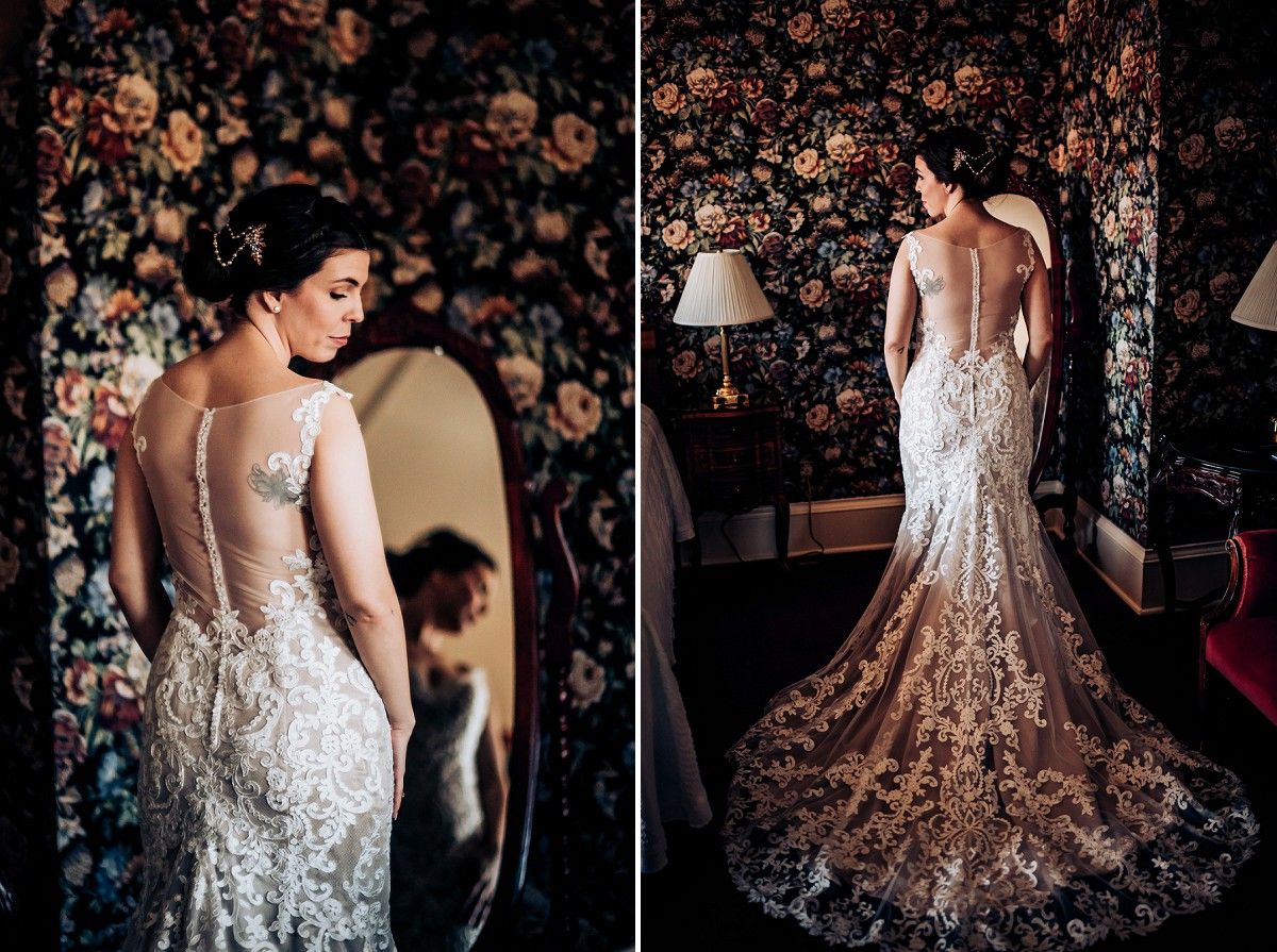 Back view of an intricate lace wedding gown against floral wallpaper in romantic lighting.