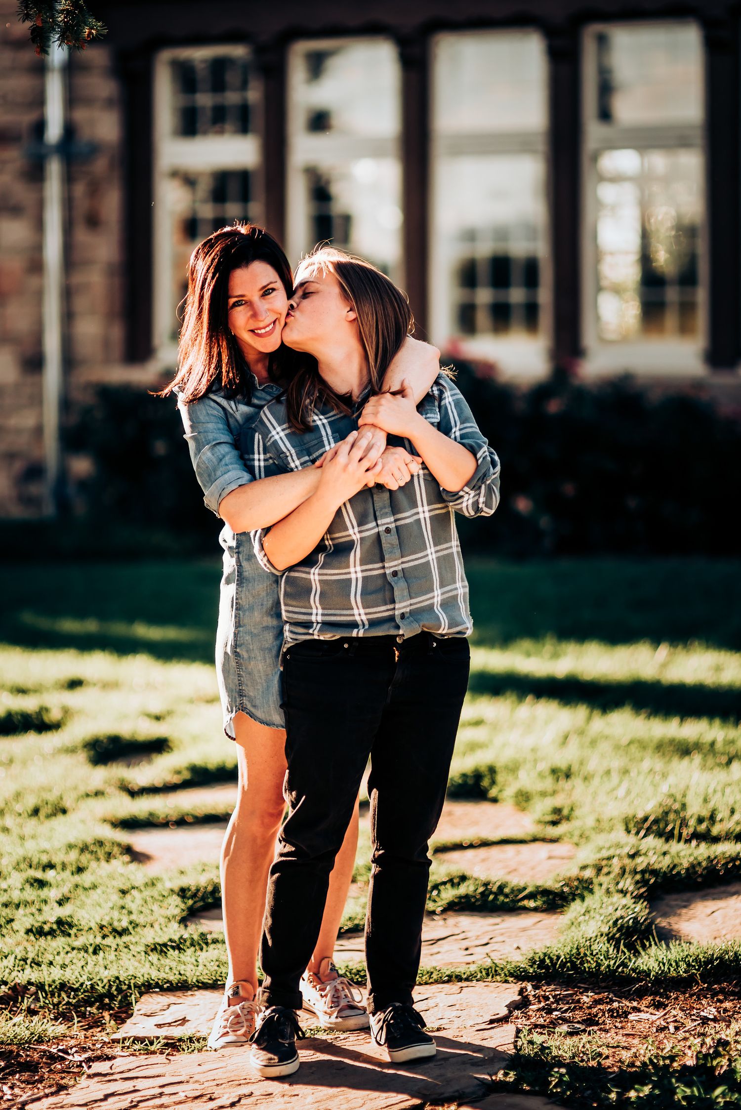 Two figures embrace outdoors with sunlit windows in the background on a sunny afternoon.