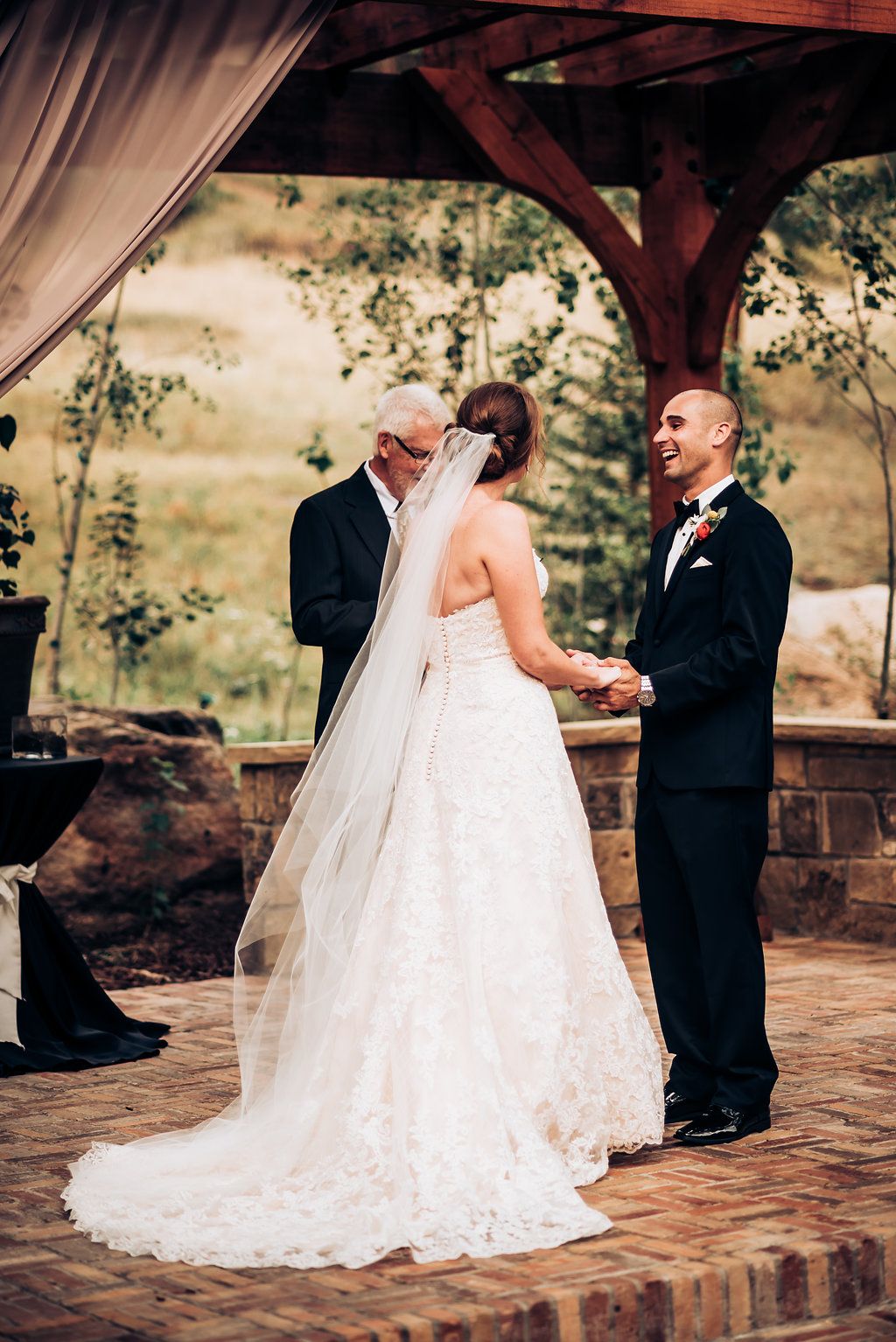 Wedding ceremony under wooden pergola with flowing white fabric and stone base details.