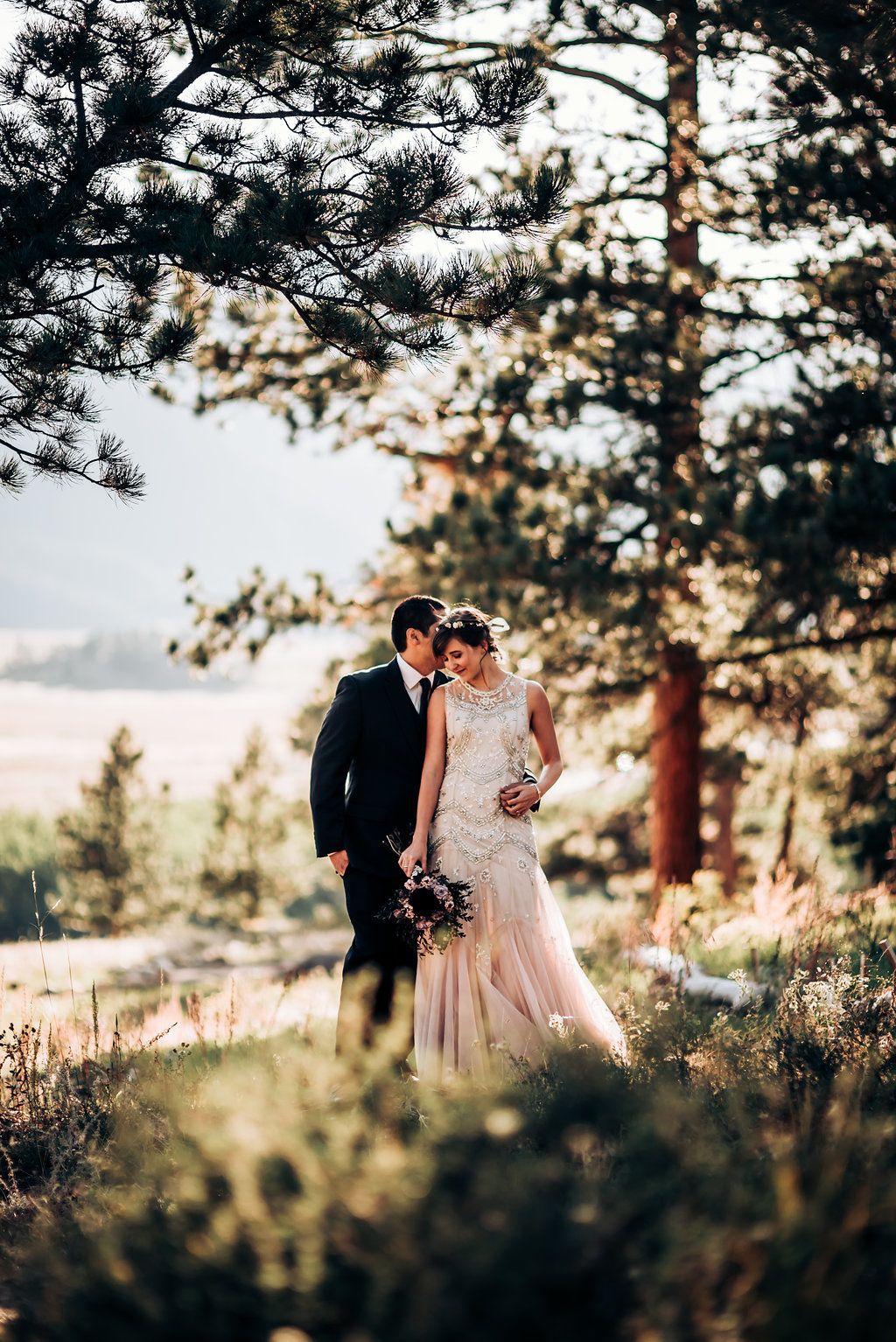A romantic wedding couple stands together among pine trees at sunset in a mountainous outdoor setting.