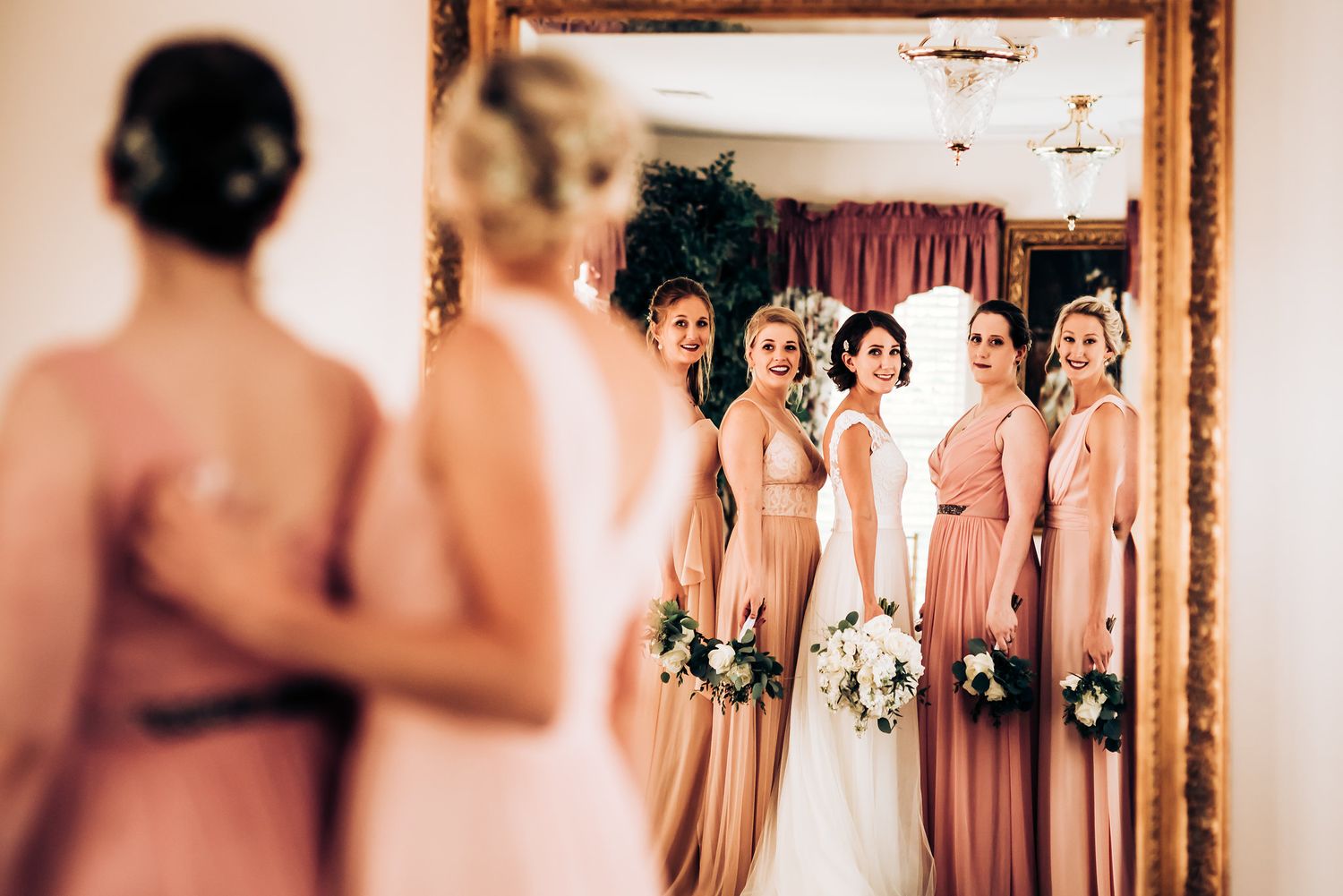 Bridesmaids in blush dresses admire the reflection in an ornate mirror.