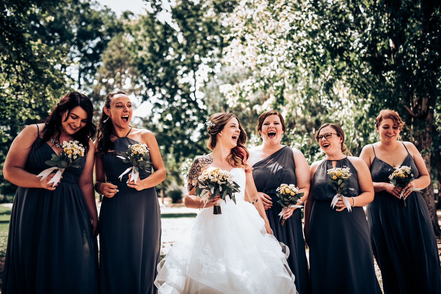 Wedding party in navy dresses poses outdoors with trees in background.