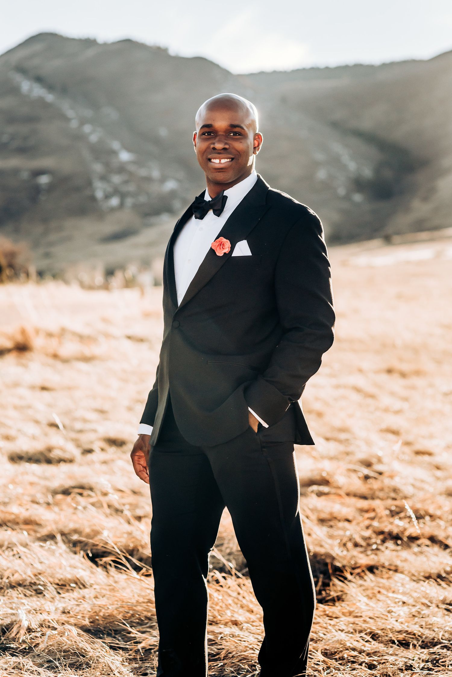 Classic groom portrait in black tuxedo smiling against mountainous landscape.