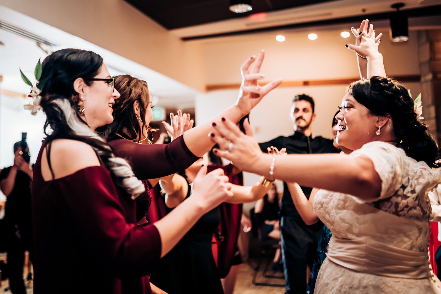 Wedding guests dance enthusiastically together with raised hands at an indoor reception celebration.