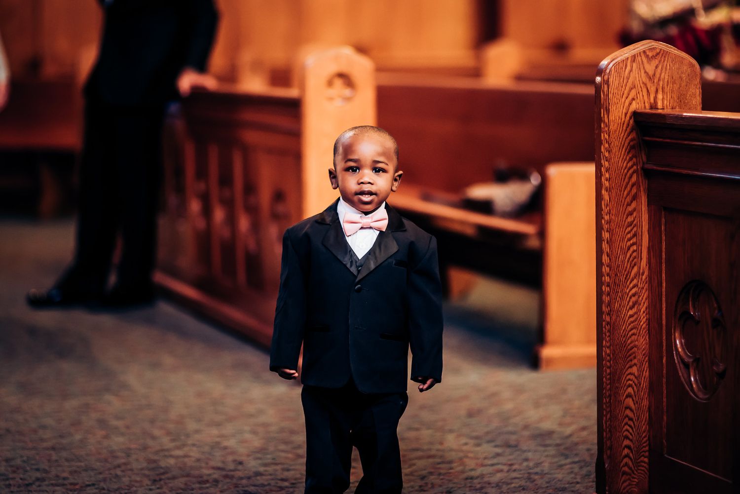 Young child in formal suit stands in church aisle with warm lighting.