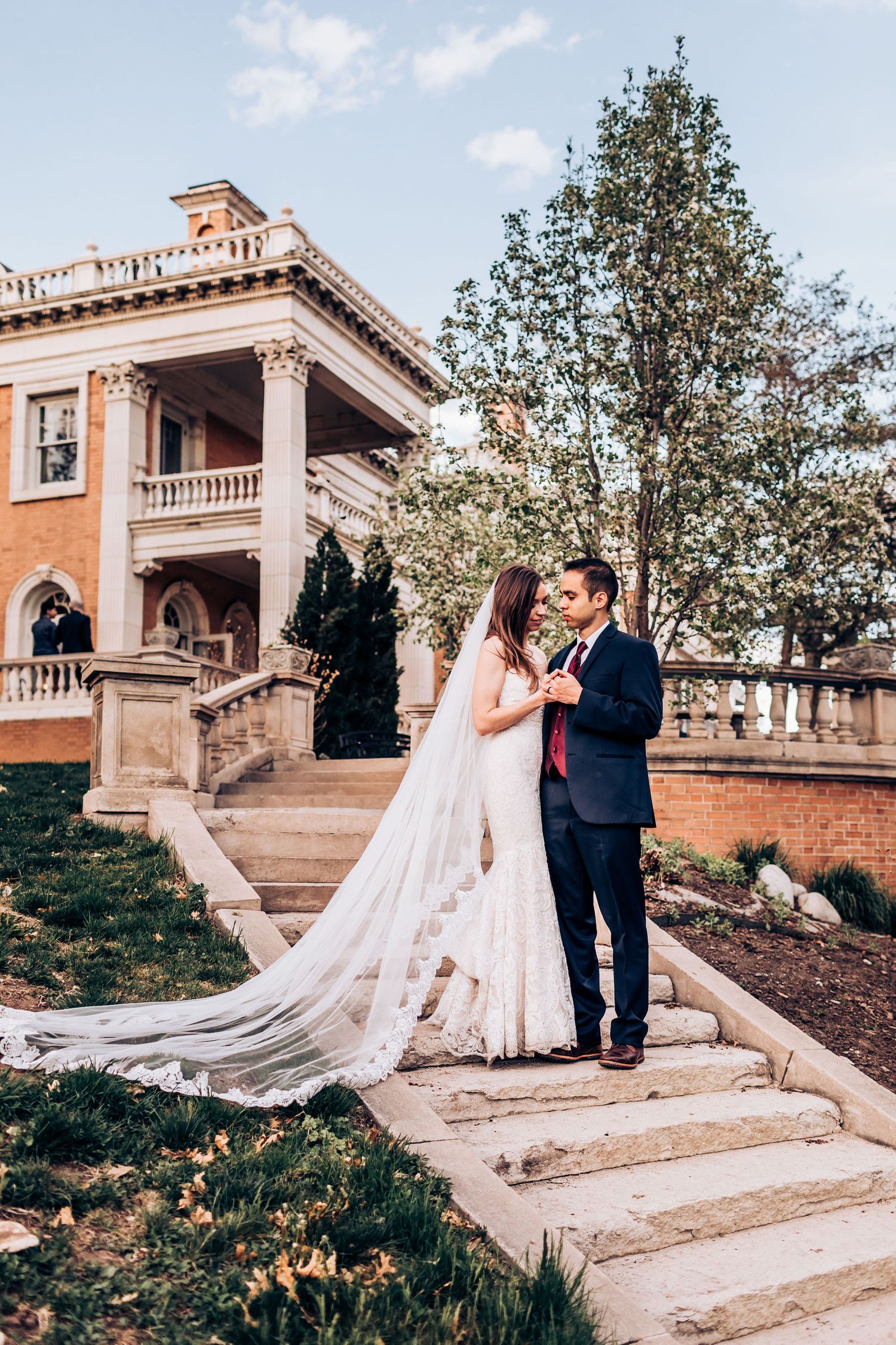 Wedding couple embraces on grand staircase of historic brick mansion with flowing cathedral veil.