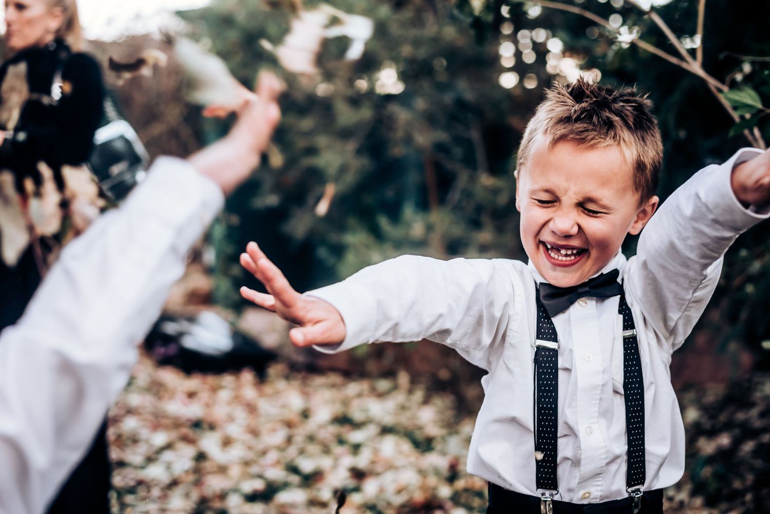 A joyful young attendant in suspenders and bowtie laughs and dances at an outdoor celebration.