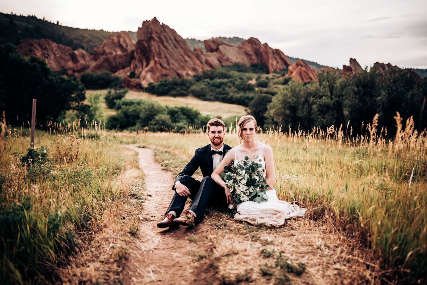 A wedding couple sits on a dirt path with red rock formations and mountains in the background at sunset.