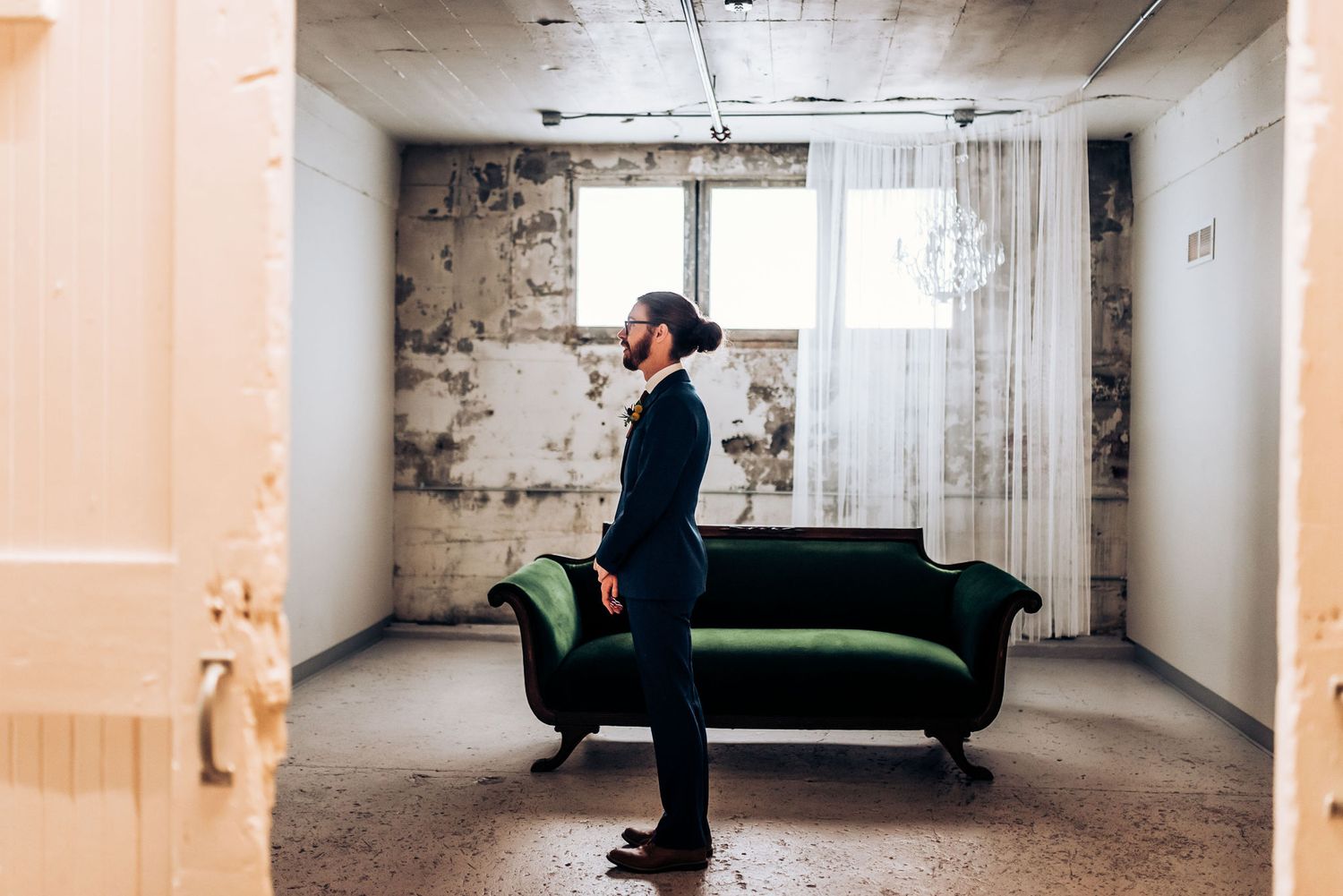 Person in dark suit stands pensively near green velvet sofa in industrial-style room.