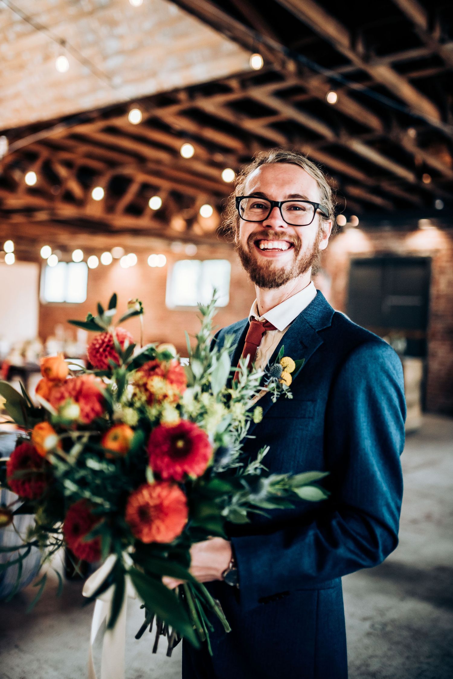 Wedding party member in navy suit and glasses holds vibrant floral bouquet in rustic venue with string lights.