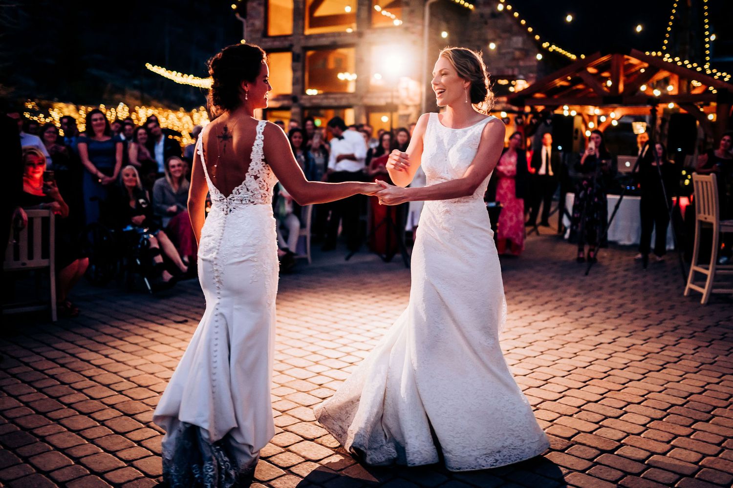 Two people in white wedding gowns dancing together on a brick patio during evening reception.