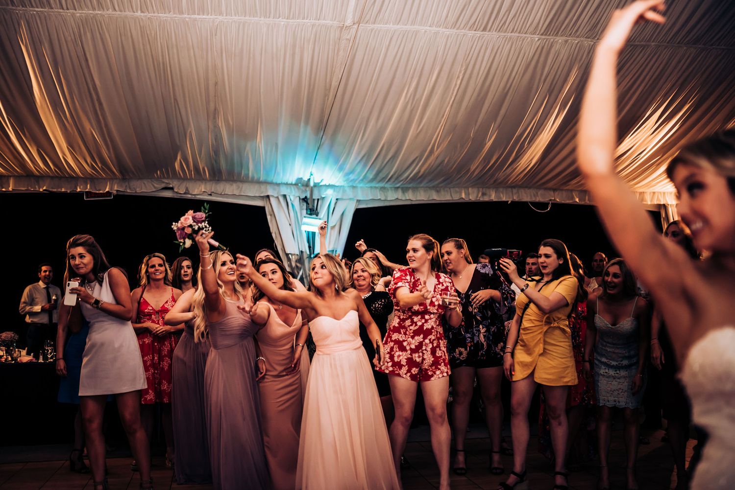 Wedding party and guests dancing together under twinkling tent lights at reception.