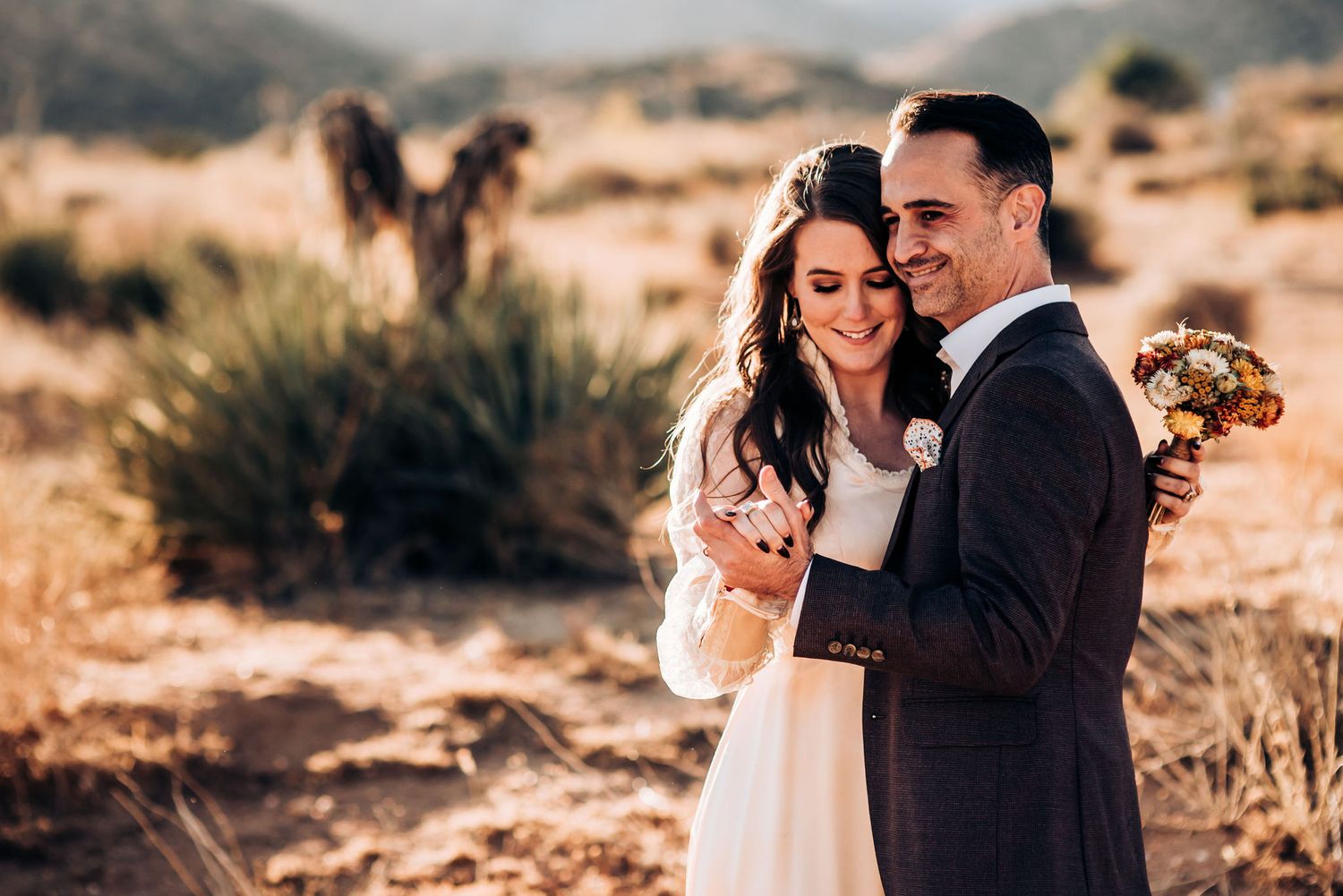 A couple shares coffee and laughter in a desert setting with mountains behind them.