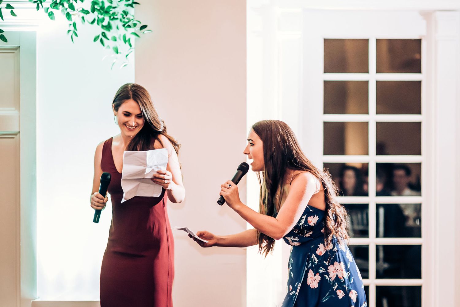 Two people sharing speaking duties at formal event with decorative greenery and white walls.