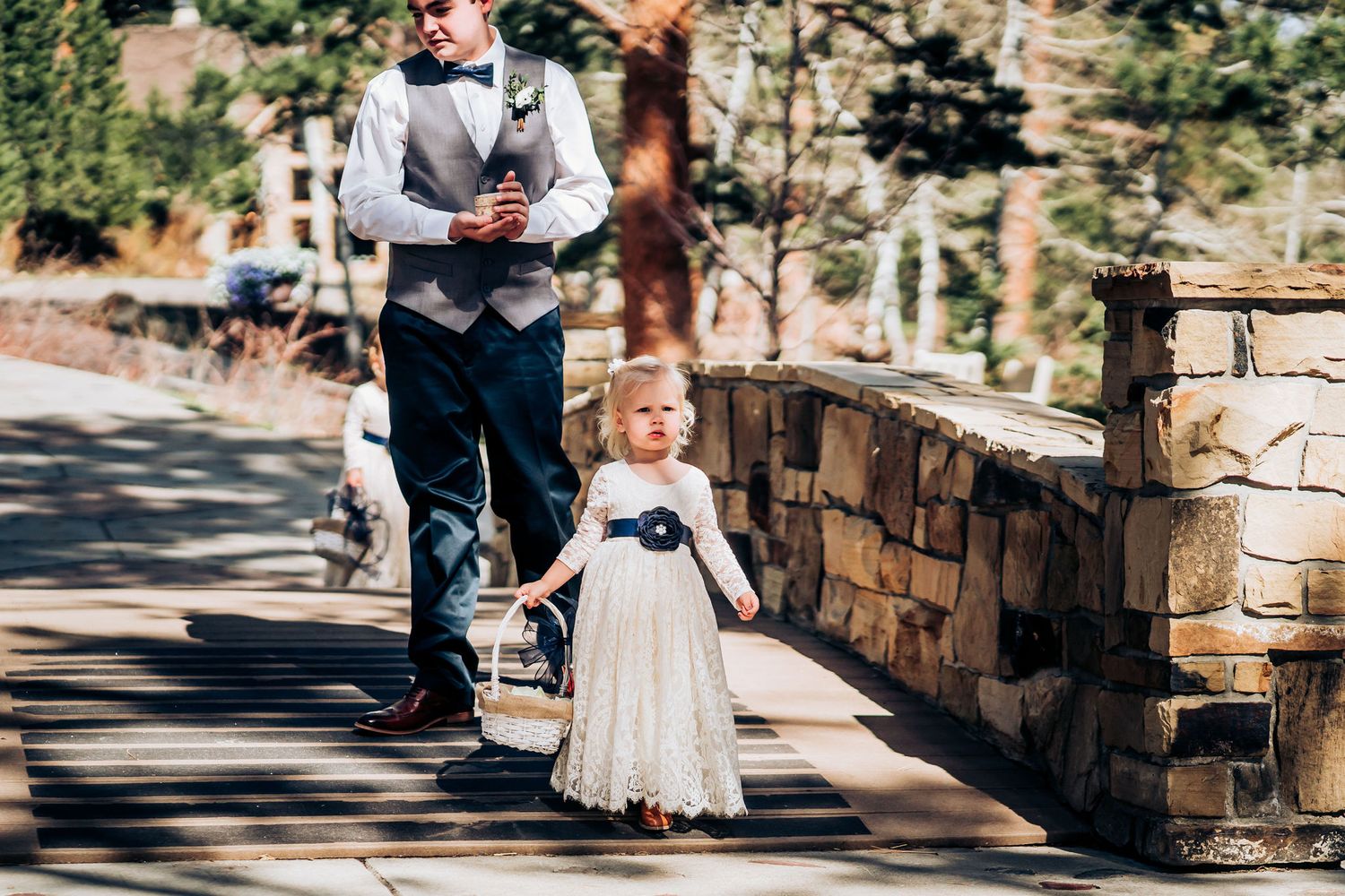 Young flower girl in white dress walks down stone-lined pathway at outdoor wedding.