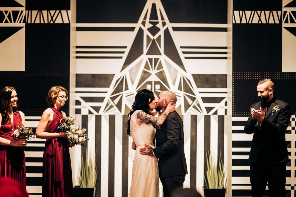 Wedding couple shares intimate moment at altar with geometric black and white backdrop and burgundy-clad wedding party.