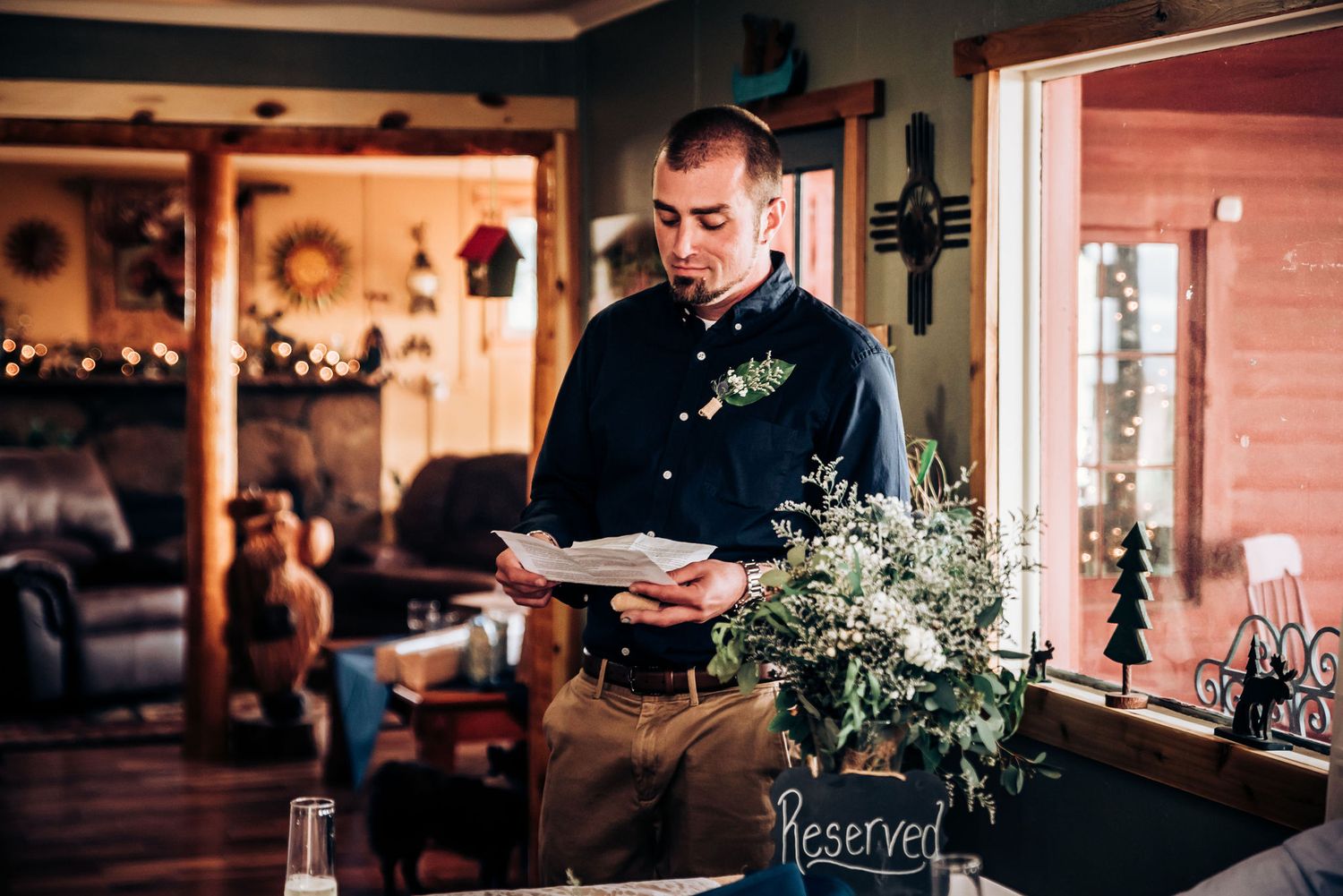 A man in black attire reads from papers while standing at a decorated wedding reception venue.