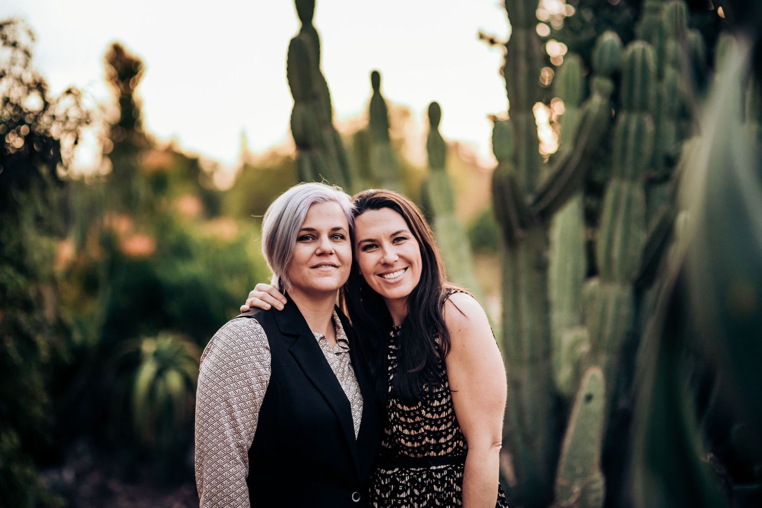 Two people embrace while standing among desert cacti at sunset.