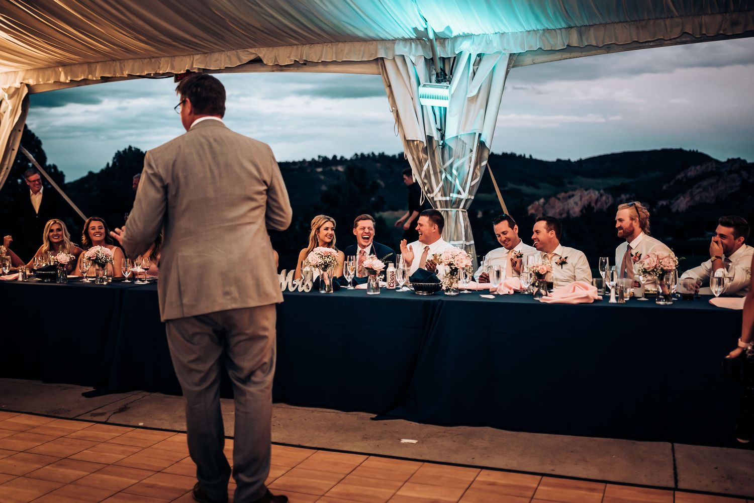 Speaker addressing wedding party at head table during outdoor reception.