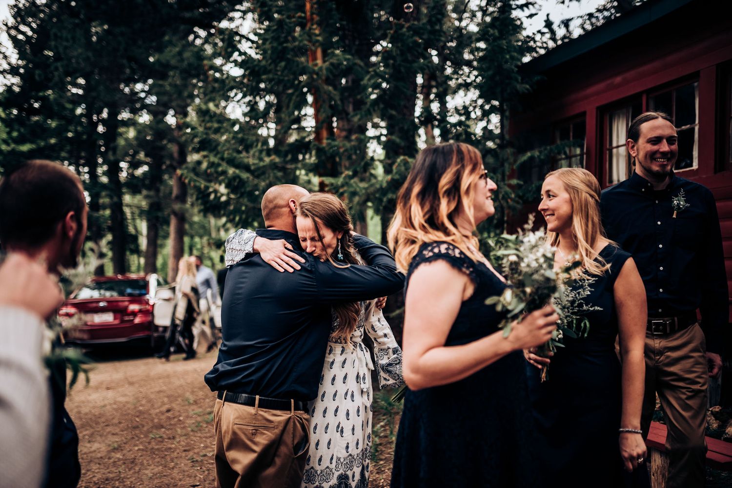 Celebratory group gathering with bouquets outside a rustic red cabin in a forest setting.