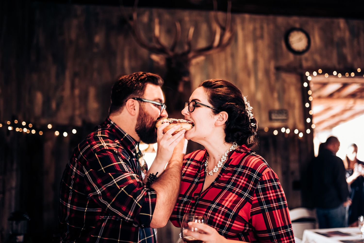 Two people in plaid sharing food in a rustic venue with string lights and antler decor.