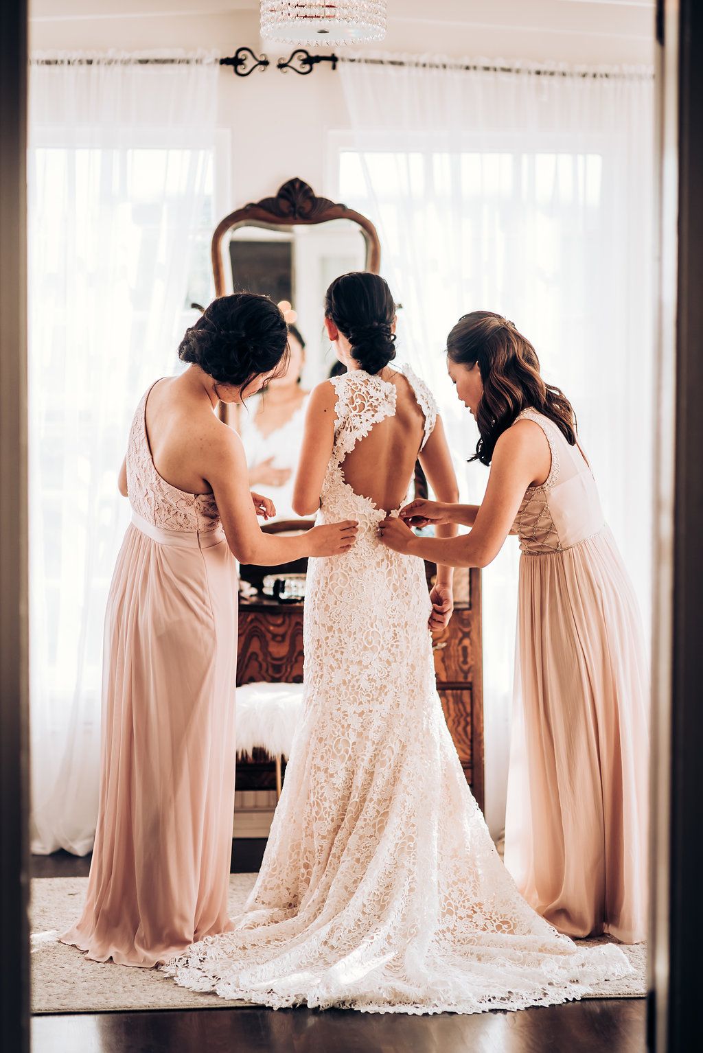 Three bridesmaids in blush pink dresses helping with a lace wedding gown in front of a vintage mirror.
