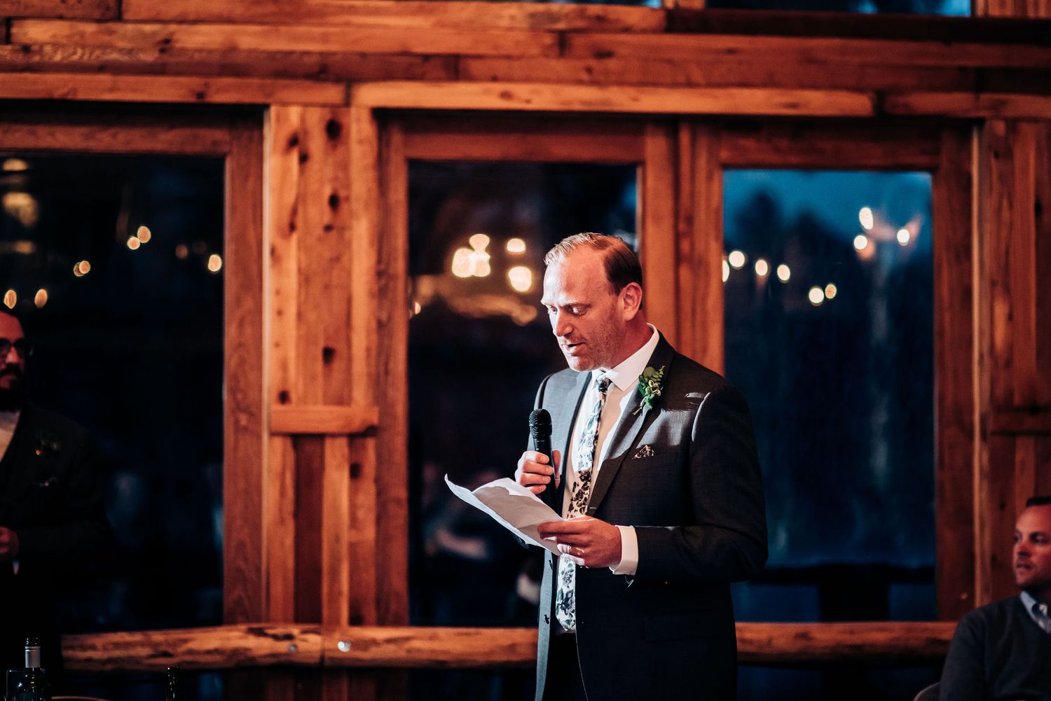 Speaker delivers remarks at evening wedding reception in wooden lodge.